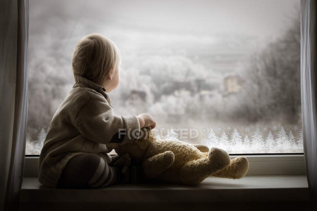 Мальчик с плюшевым мишкой и смотрящим в окно — стоковое фото