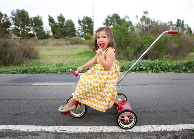 Girl on tricycle eating lollipop — Stock Photo