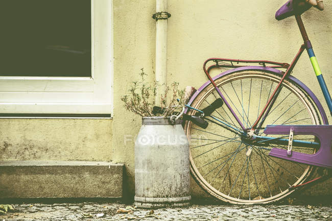 Vélo enchaîné à l'extérieur d'une maison à côté d'un seau de lait avec des fleurs — Photo de stock