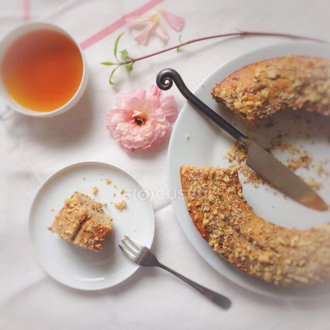 Banana bread with piece on plate and cup of tea on white tablecloth with flowers — Stock Photo