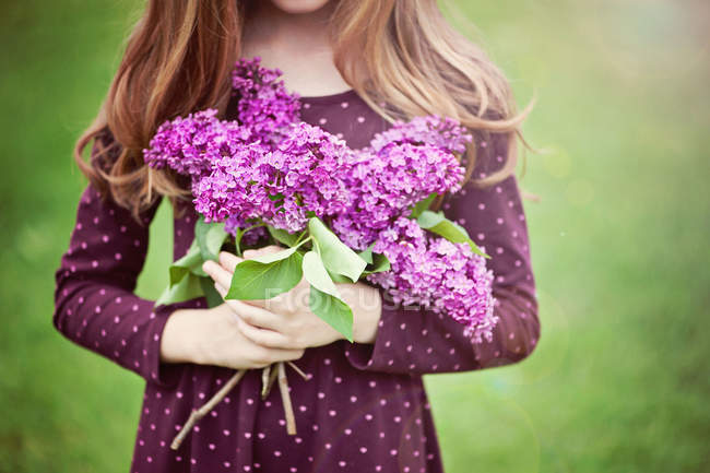 Imagen recortada de niña sosteniendo ramo de flores lila sobre fondo borroso - foto de stock