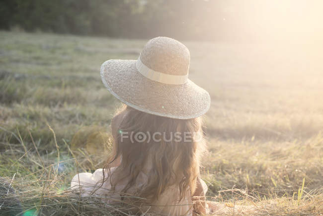 Vista posteriore di una ragazza seduta indossando cappello di paglia in campo — Foto stock