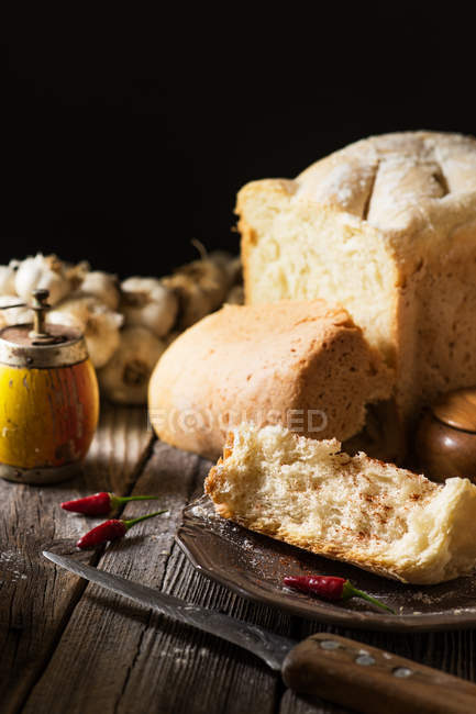 Loaf of bread, garlic and chilies on rustic wooden table against black background — Stock Photo