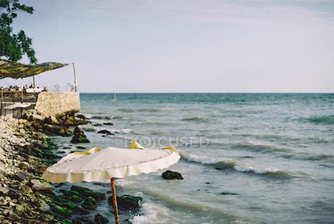 Día de verano en la playa rocosa con sombrilla blanca y gente borrosa en el fondo - foto de stock