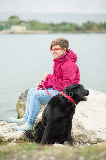 Sonriente joven Chica sentada en el tronco cerca del lago con perro - foto de stock