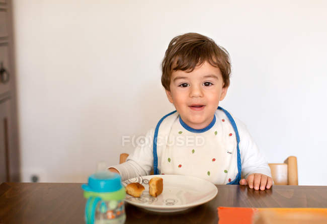 Sonriente niño sentado en la mesa comiendo bocadillo - foto de stock