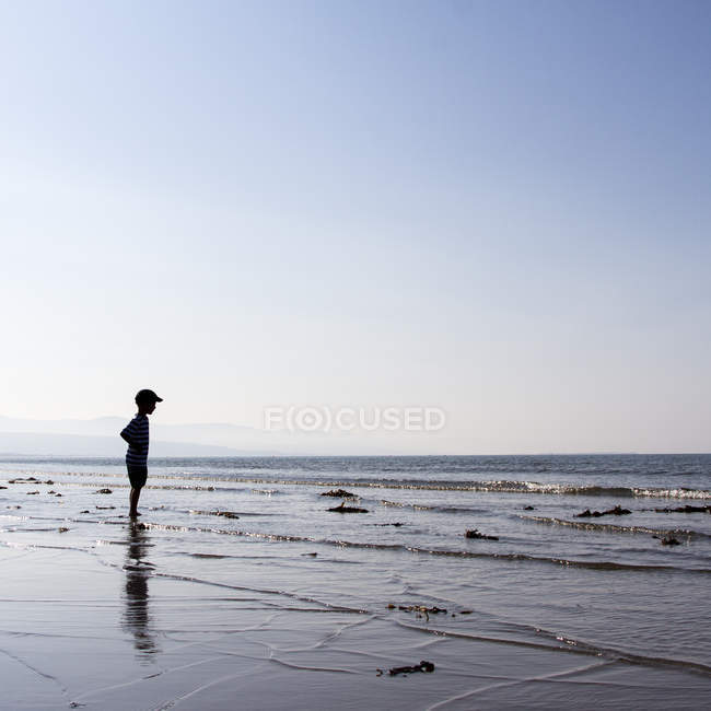 Niño de pie en la playa y mirando al mar - foto de stock