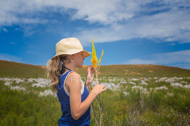 Fille portant chapeau tenant des fleurs sauvages sur prairie — Photo de stock