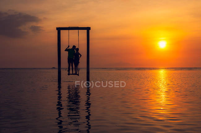 Silhueta de homem e mulher beijando, de pé em um balanço no mar ao pôr do sol — Fotografia de Stock