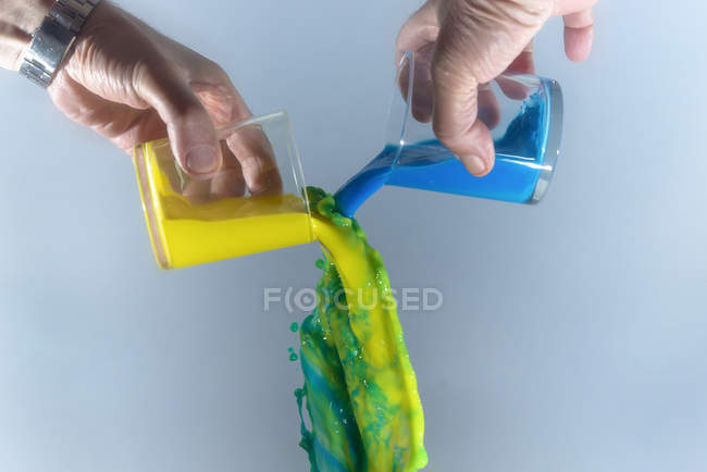 Чоловічі руки Змішування жовтої та синьої рідини, щоб отримати зелену рідину на сірому фоні — стокове фото