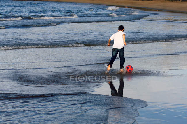 Garçon coups de pied football dans l'eau sur la plage — Photo de stock
