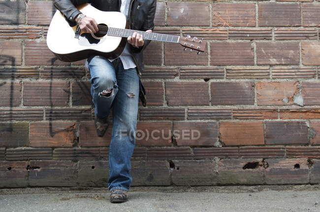 Обрезанное изображение человека в кожаной куртке и ковбойских сапогах, играющего на гитаре в переулке — стоковое фото