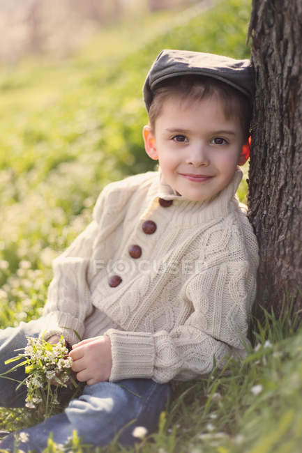 Boy leaning against a tree trunk, holding bunch of spring flowers — Stock Photo