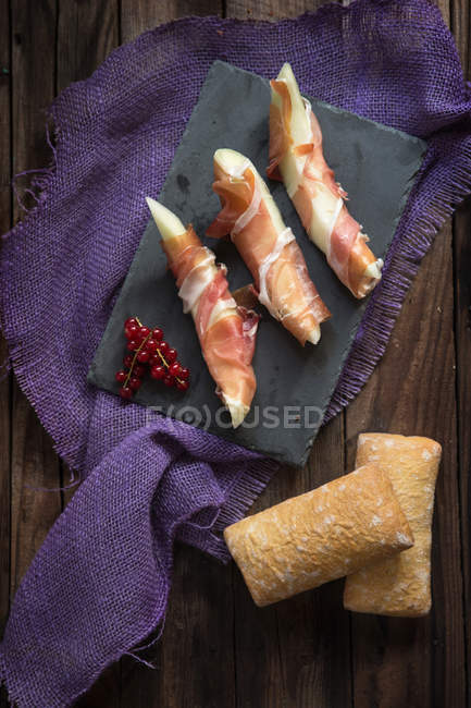 Melon wrapped in parma ham with bread on slate — Stock Photo