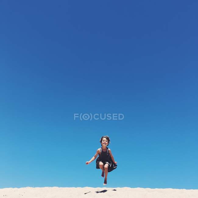Adorable little girl jumping on sandy beach against blue sky — Stock Photo