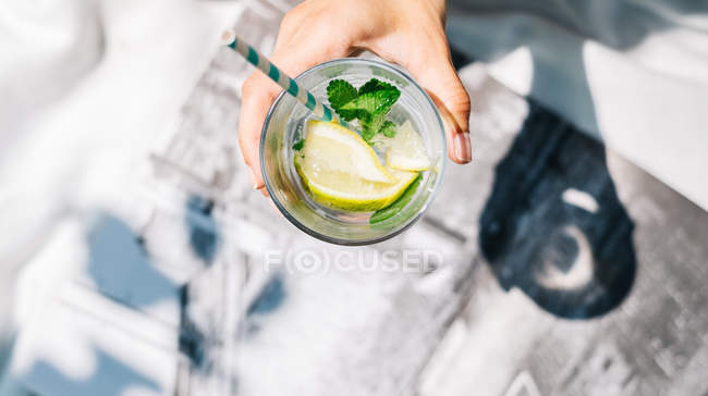 Overhead view of hand holding glass of water with fresh lemon, lime, mint and ice-cubes — Stock Photo