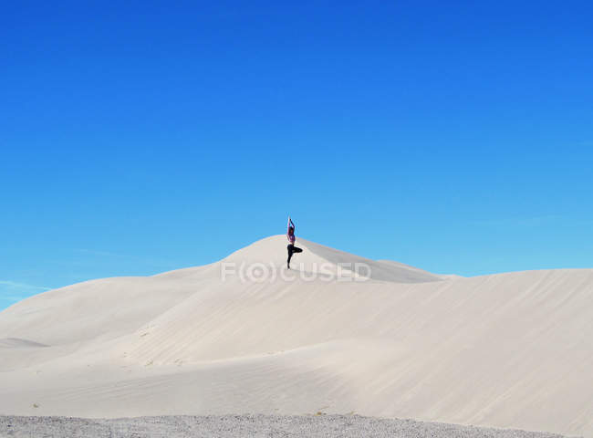 Женщина, занимающаяся йогой, позирует на песчаной дюне на фоне голубого неба — стоковое фото