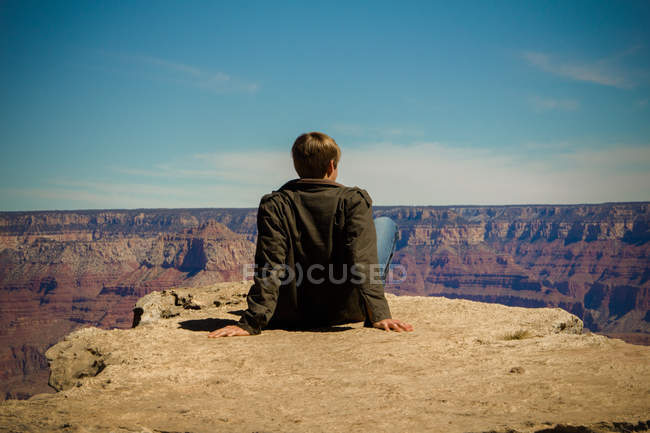 Вид сзади на молодого человека, сидящего на краю большого каньона и смотрящего на вид, Аризона, США — стоковое фото