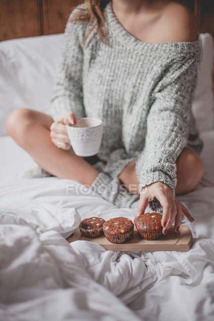 Donna seduta sul letto con tazza di tè e muffin — Foto stock