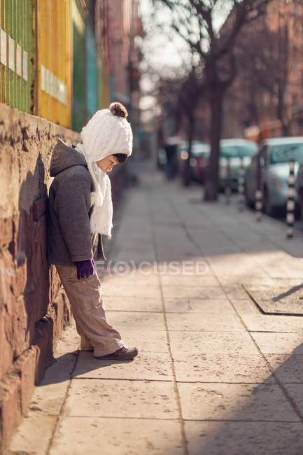 Сумний хлопчик спирається на стіну, дивлячись вниз на тротуар — стокове фото