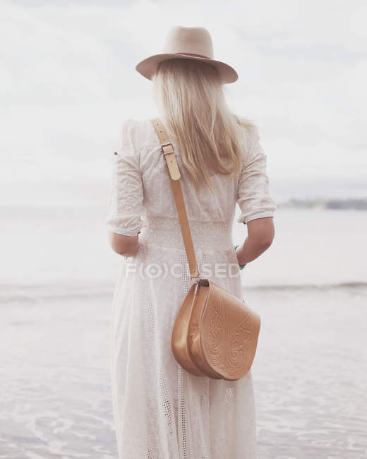 Vista trasera de la mujer de pie en la playa con vestido blanco y sombrero - foto de stock
