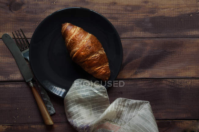 Верхний вид круассана на тарелку, салфетку и кухонную утварь на деревянный стол — стоковое фото