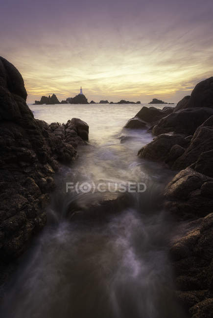 Джерси, Ла-Корбьер, морская вода, текущая между скалами с маяком, видимым на расстоянии — стоковое фото