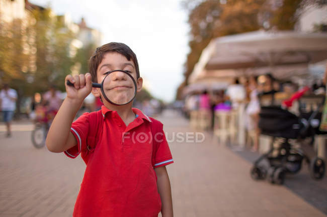 Boy smiling through magnifying glass on street — Stock Photo
