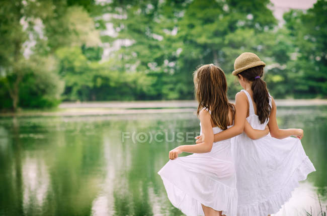 Rear view of two cute sisters standing in white dresses at lake in forest — Stock Photo