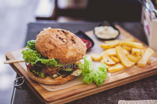Hambúrguer saboroso com batatas fritas e molho em uma placa de madeira — Fotografia de Stock