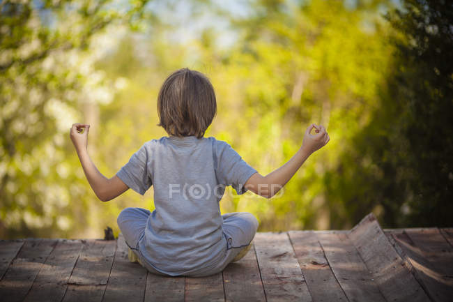 Vue arrière d'un garçon assis sur une terrasse en bois et méditant — Photo de stock