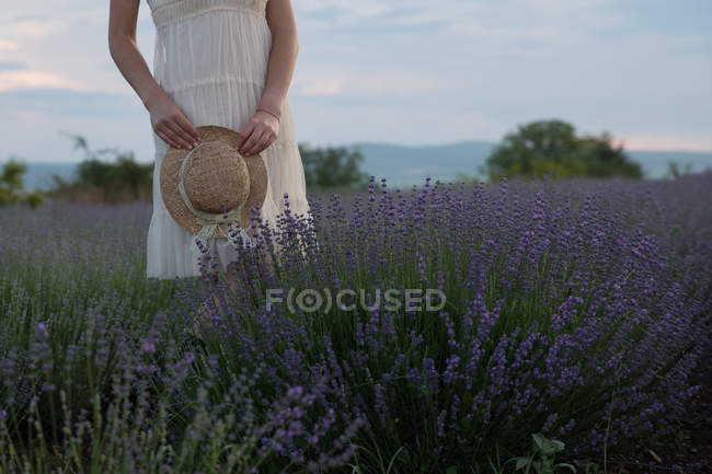 Обрезанное изображение женщины, стоящей на лавандовом поле и держащей соломенную шляпу — стоковое фото