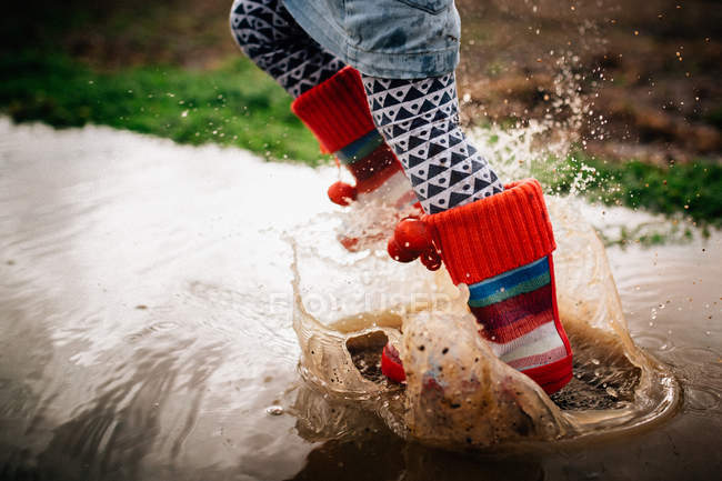 Imagem cortada de pernas de criança correndo em poça enlameada em botas coloridas — Fotografia de Stock