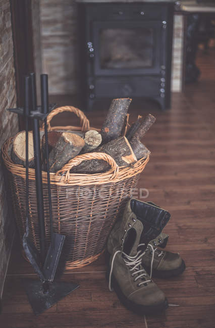Boots next to a basket filled with firewood and hearth set — Stock Photo