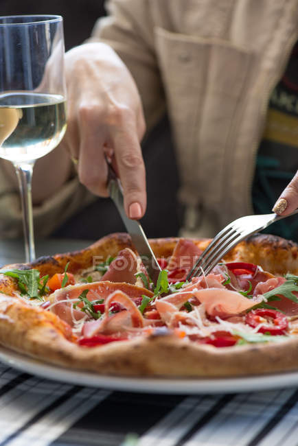 Mãos femininas cortando um pedaço de pizza deliciosa e um copo de vinho branco na mesa — Fotografia de Stock
