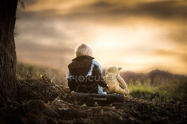 Vista posteriore del ragazzo seduto sulle radici dell'albero con orsacchiotto — Foto stock