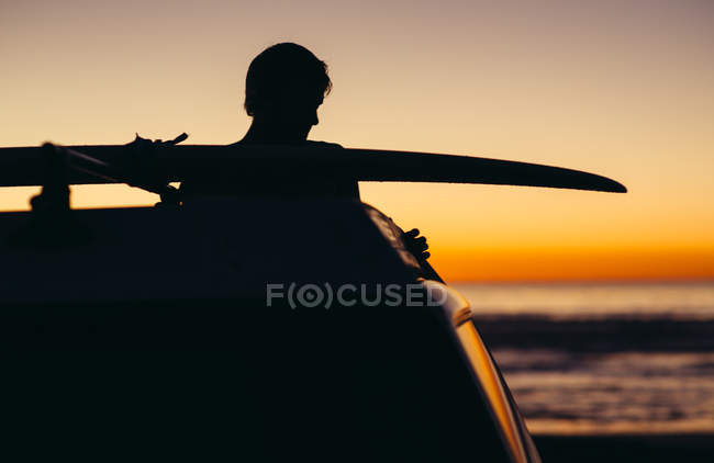Силует автомобіля і молода людина з дошки для серфінгу на даху проти гарний захід сонця в Сан-Дієго, Америка, США — стокове фото