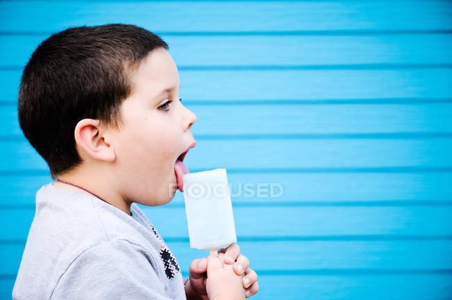 Menino lambendo gelo alegre na frente da parede azul — Fotografia de Stock