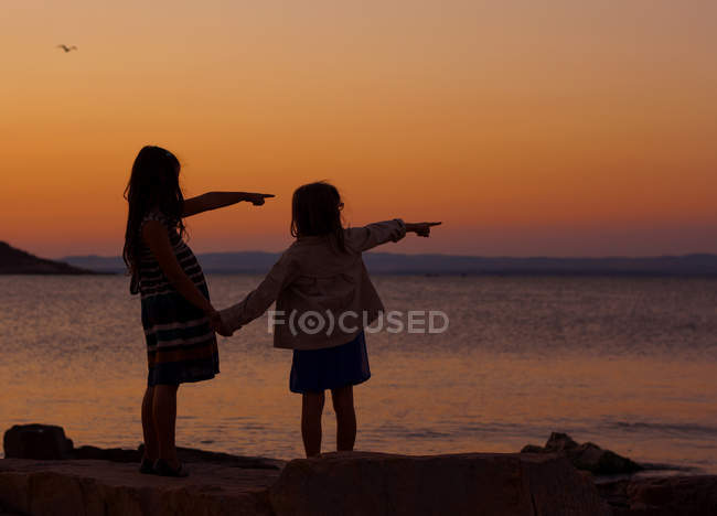 Силует двох молодих дівчат, що вказують на море на заході сонця — стокове фото