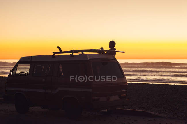 Homem colocando prancha na prateleira do telhado ao pôr do sol na praia — Fotografia de Stock