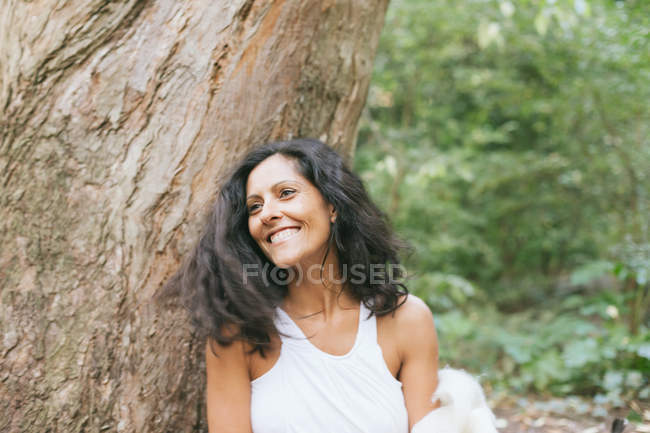 Портрет улыбающейся женщины, прислонившейся к дереву в парке — стоковое фото