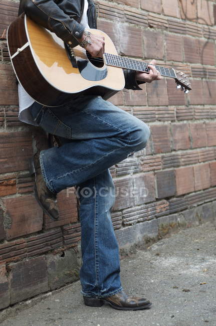 Abgeschnittenes Bild eines Mannes in Lederjacke und Cowboystiefeln, der in einer Gasse Gitarre spielt — Stockfoto