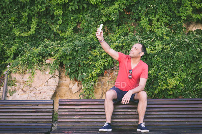 Portrait of a man sitting on bench and taking selfie with smartphone — Stock Photo
