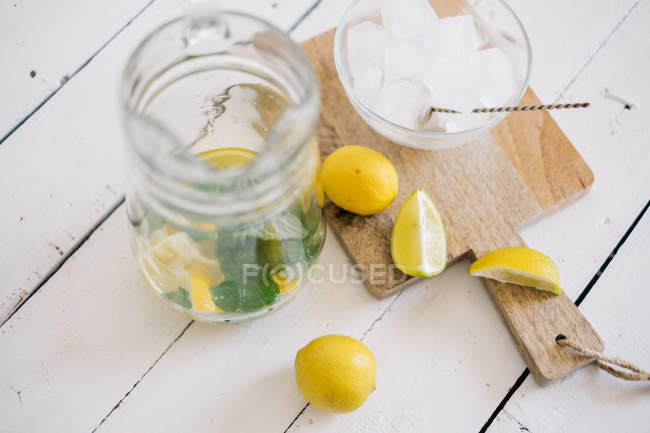 Jarro de vidro e tábua de cortar com limão fresco, limão, hortelã e cubos de gelo — Fotografia de Stock