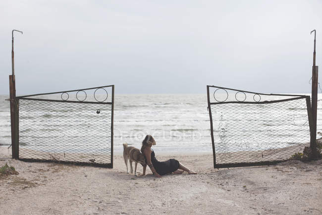 Vista lateral de la joven sentada en la playa de arena con perro - foto de stock
