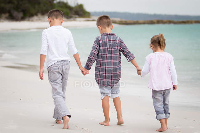 Rückansicht glücklicher Kinder, die am Strand spazieren und Händchen halten — Stockfoto