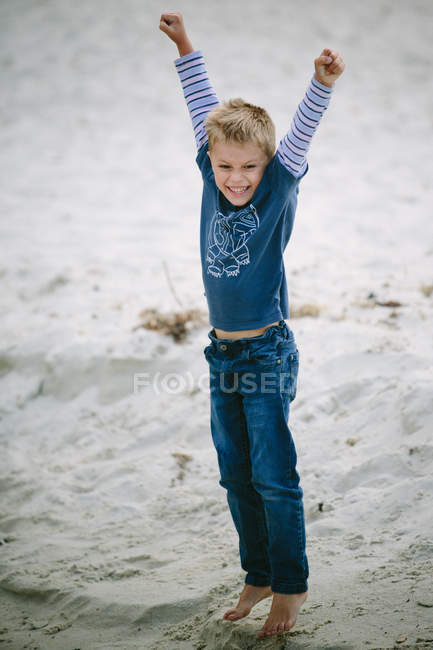 Niño feliz de pie en la playa de arena con los brazos levantados - foto de stock