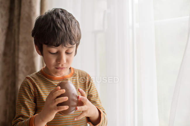 Мальчик стоит у окна и ест большое шоколадное пасхальное яйцо — стоковое фото