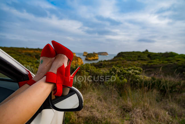 Женщина отдыхает на ногах в красных туфлях на каблуках на двери машины через открытое окно — стоковое фото