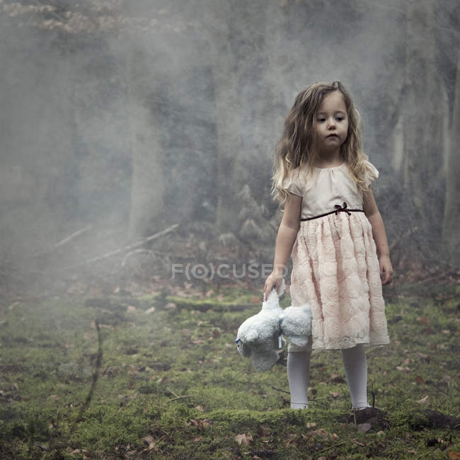 Girl wearing dress standing in woods and holding a teddy bear — Stock Photo